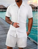 A stylish man with a muscular build is wearing a short-sleeved, button-up green shirt with matching green shorts. He is accessorized with silver rings, a wristwatch, and a layered necklace. His left arm is tattooed, and he is posing casually near a modern poolside setting with a blurred outdoor background.