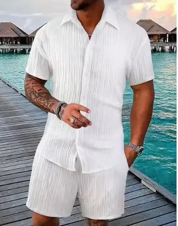 A stylish man with a muscular build is wearing a short-sleeved, button-up green shirt with matching green shorts. He is accessorized with silver rings, a wristwatch, and a layered necklace. His left arm is tattooed, and he is posing casually near a modern poolside setting with a blurred outdoor background.