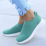A pair of beige slip-on knit sneakers with a lightweight sock-like design and white flexible soles. The shoes feature breathable mesh texture, ribbed detailing across the front, and are shown worn with light blue distressed jeans.