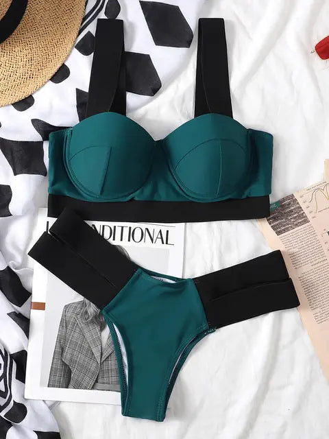 Teal bikini set with molded underwire top and high-cut bottoms, both with wide black elastic straps, laid flat on a patterned bedspread with magazines and a straw hat nearby.