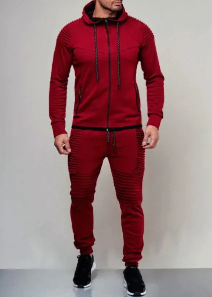 A man is wearing a stylish, deep red tracksuit with a fitted design. The hoodie features a front zipper, ribbed detailing on the shoulders, and black drawstrings. The matching joggers have ribbed accents on the thighs and knees, with black drawstrings at the waist. He completes the outfit with black sneakers that have white soles. The background is a simple, neutral-colored wall, emphasizing the bold red of the tracksuit.