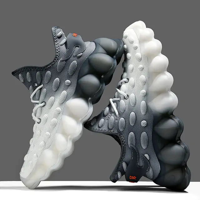A pair of futuristic gray-and-white athletic sneakers with a textured knit upper, lace-up front, and exaggerated bubble-like sculpted soles. The shoes feature a gradient color fade, raised oval detailing across the upper, and a bold high-concept design that gives them a striking, modern look.