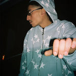 Person wearing a light gray zip-up hoodie with white star print, hood up, and glasses, photographed in a dark setting with one hand extended forward.