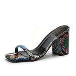 Open-toe mule sandal with a clear upper strap and a multicolor snakeskin-pattern block heel and toe strap, on a black footbed.