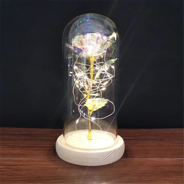 A decorative tabletop lamp featuring a rose inside a clear glass dome (cloche) on a light wooden base, wrapped with warm white string/fairy lights that glow against a dark background, sitting on a wooden surface.