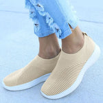 Person wearing beige slip-on knit sneakers with white ribbed soles, paired with light blue distressed jeans, standing on a concrete surface.