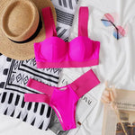Flat lay of a bright hot pink bikini set with a structured top and high-cut bottoms with wide side straps, styled on a bed with a straw hat, patterned towel, pink-tinted sunglasses, and an open magazine.
