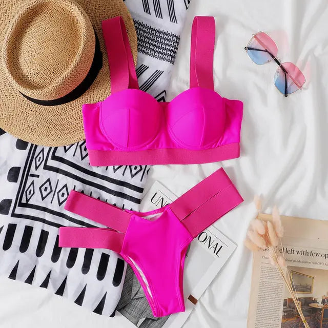 Flat lay of a bright hot pink bikini set with a structured top and high-cut bottoms with wide side straps, styled on a bed with a straw hat, patterned towel, pink-tinted sunglasses, and an open magazine.