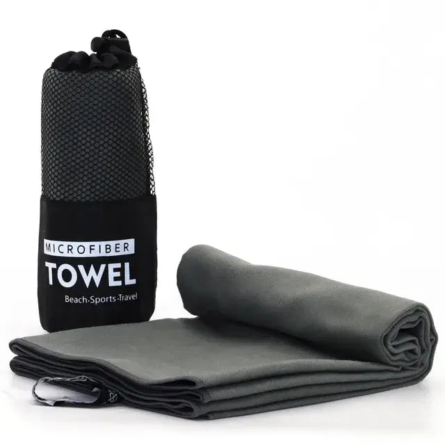 Gray microfiber towel folded and rolled beside a black mesh carry bag labeled ‘MICROFIBER TOWEL’ with the words ‘Beach-Sports-Travel,’ shown on a white background.