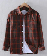 A brown and red plaid flannel shirt displayed on a wooden hanger against a neutral background. The shirt features a button-up front, a structured collar, long sleeves with buttoned cuffs, and a single chest pocket. It is layered over a plain white T-shirt, giving it a casual and stylish look. The slightly open front and draped fabric create a relaxed aesthetic.