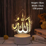 A warm glowing acrylic LED night light featuring Arabic calligraphy for “Allah” on a white round base. The lamp is shown on a tabletop against a wood-panel background, with nearby books and a small teddy bear, and includes text indicating USB power with dimensions of 18 cm height and 10 cm width.