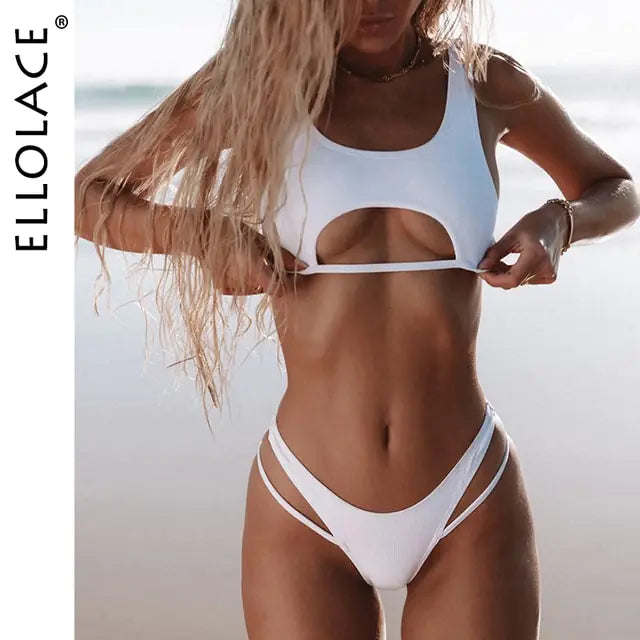 A beach lifestyle photo of a woman wearing a white two-piece bikini. The top is a sporty, tank-style cut with a front cutout under the bust. The bottoms are high-cut with double side straps. The model has long, wavy blonde hair, and the ocean shoreline is blurred in the background. The brand text “ELLOLACE” appears vertically along the left side.